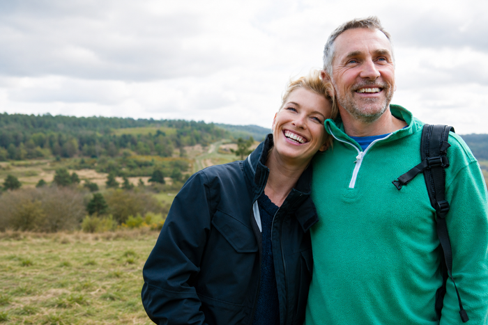 A smiling couple outdoors in a grassy field under a cloudy sky, with the man wearing a green quarter-zip fleece and the woman wearing a dark navy jacket.