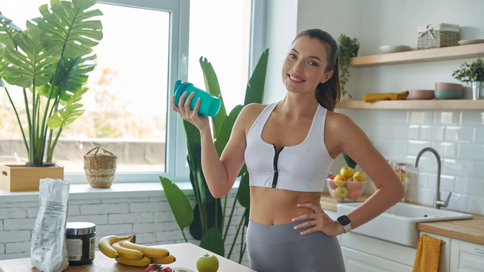A woman in a white sports bra and gray leggings holds a teal shaker bottle in a bright kitchen setting.