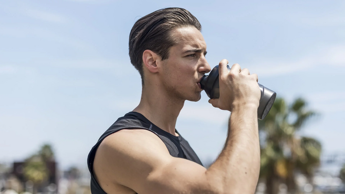A man in a black athletic tank top drinking from a dark gray shaker bottle outdoors under a clear blue sky.