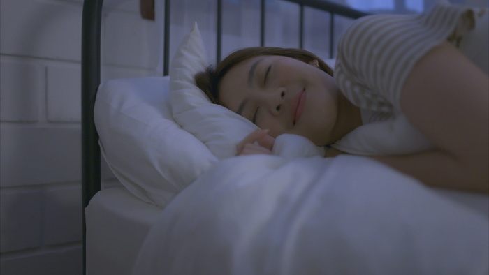 A woman sleeping soundly on a bed with white pillows and a striped shirt, captured in a cool-toned, softly lit bedroom setting.